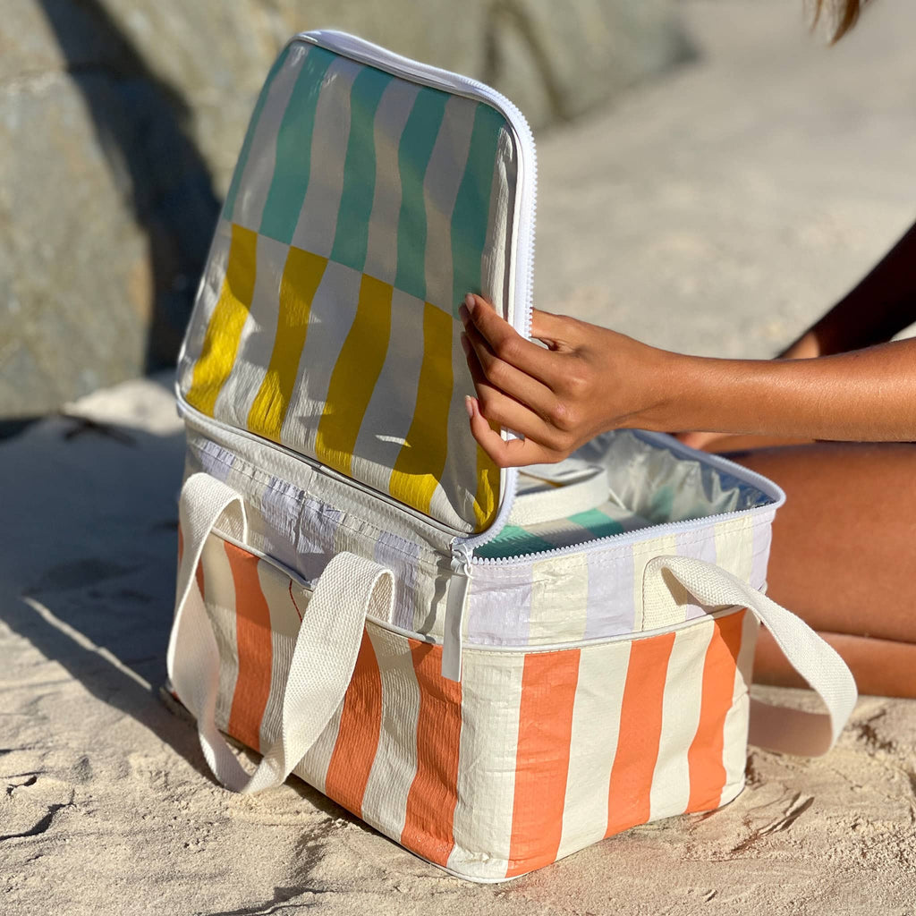 Sunnylife Cooler Bag in Rio Sun with blue, yellow, lilac, orange and cream maxi stripes, in use on a beach, lid open.