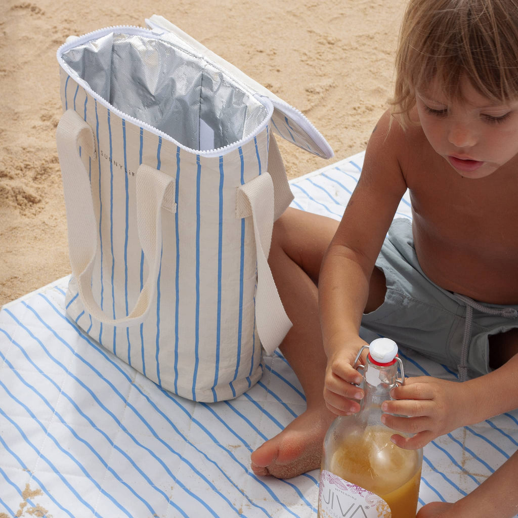 Sunnylife Insulated Drinks Cooler Bag in Le Weekend in white with blue pin stripes, on beach with lid open with child on matching beach blanket.