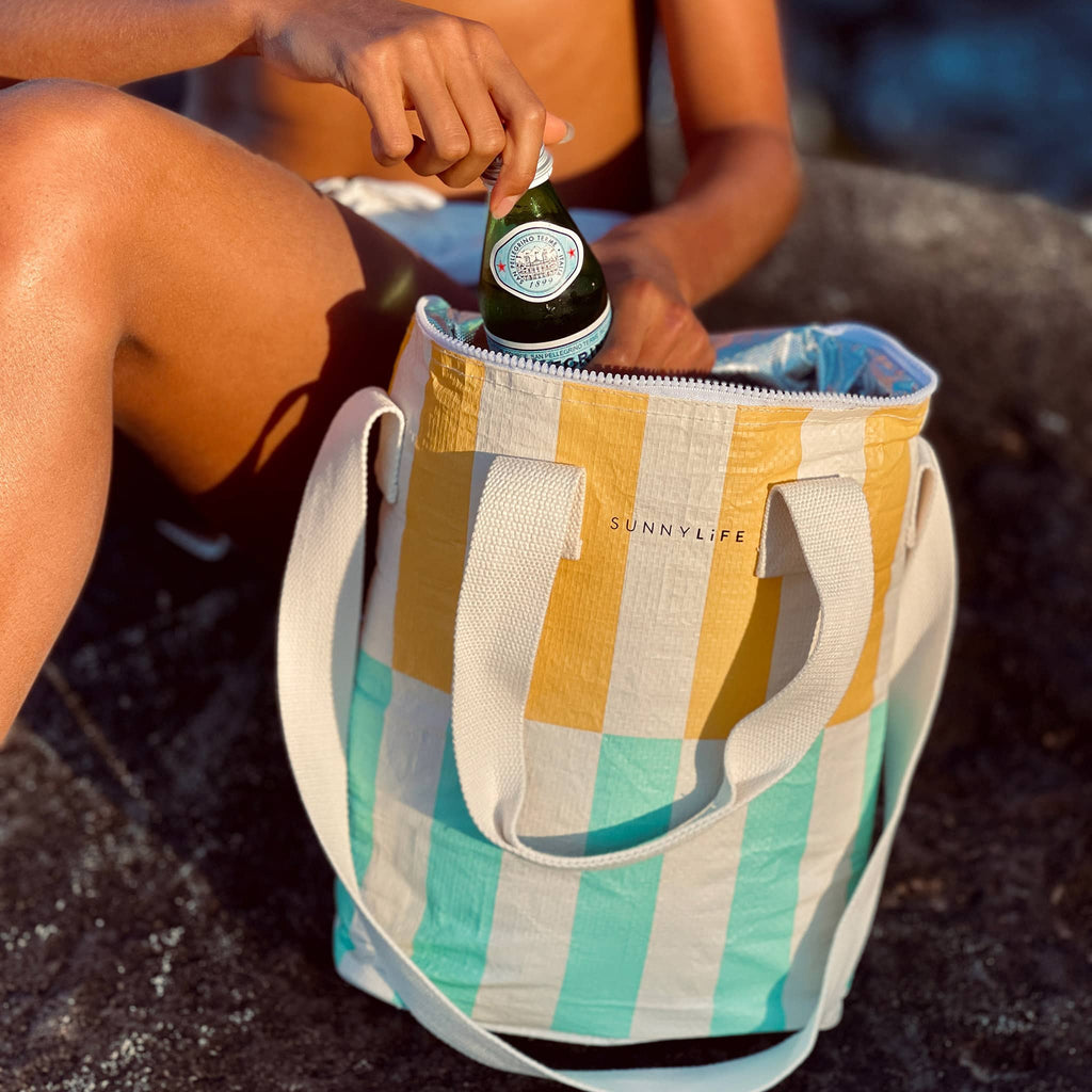 Sunnylife Insulated Drinks Cooler Bag in Rio Sun with yellow and aqua blue stripes, on the beach, lid open with a person reaching in for a bottle of sparkling water.