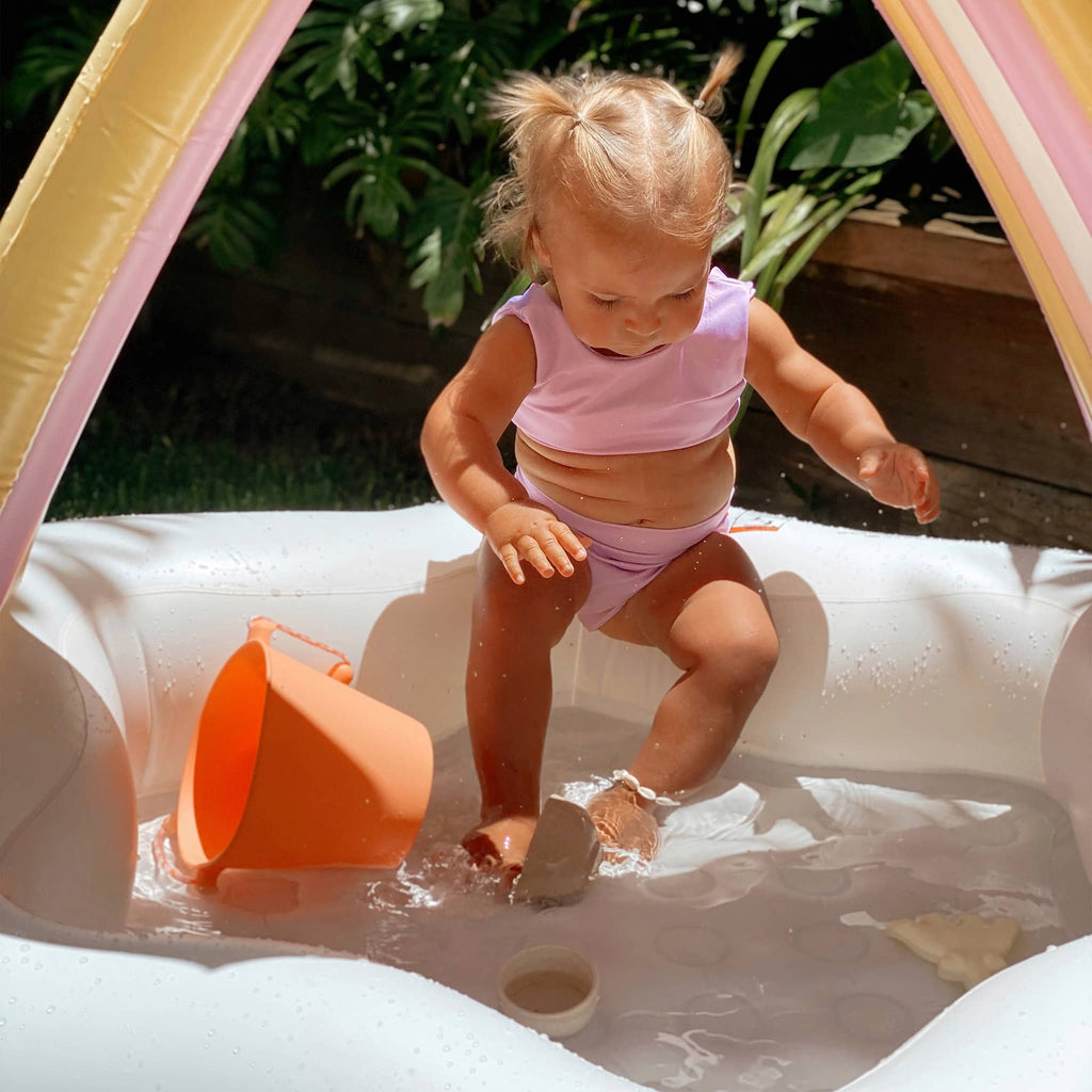 Sunnylife Princess Swan Kids Inflatable Pool with lilac, orange, pink and white tube canopy and white star-shaped pool, with a toddler.