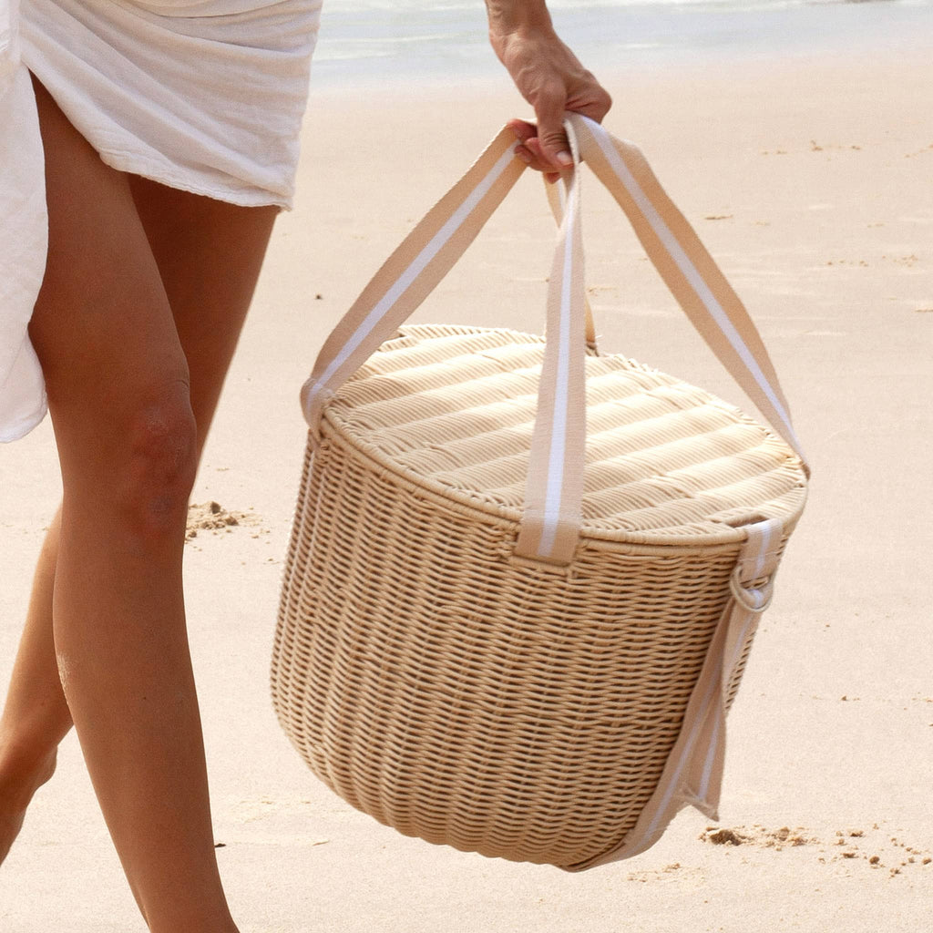 Sunnylife Round Plastic Rattan Insulated Picnic Cooler Basket in Le Weekend with natural and white webbed straps, on the beach with model carrying it.