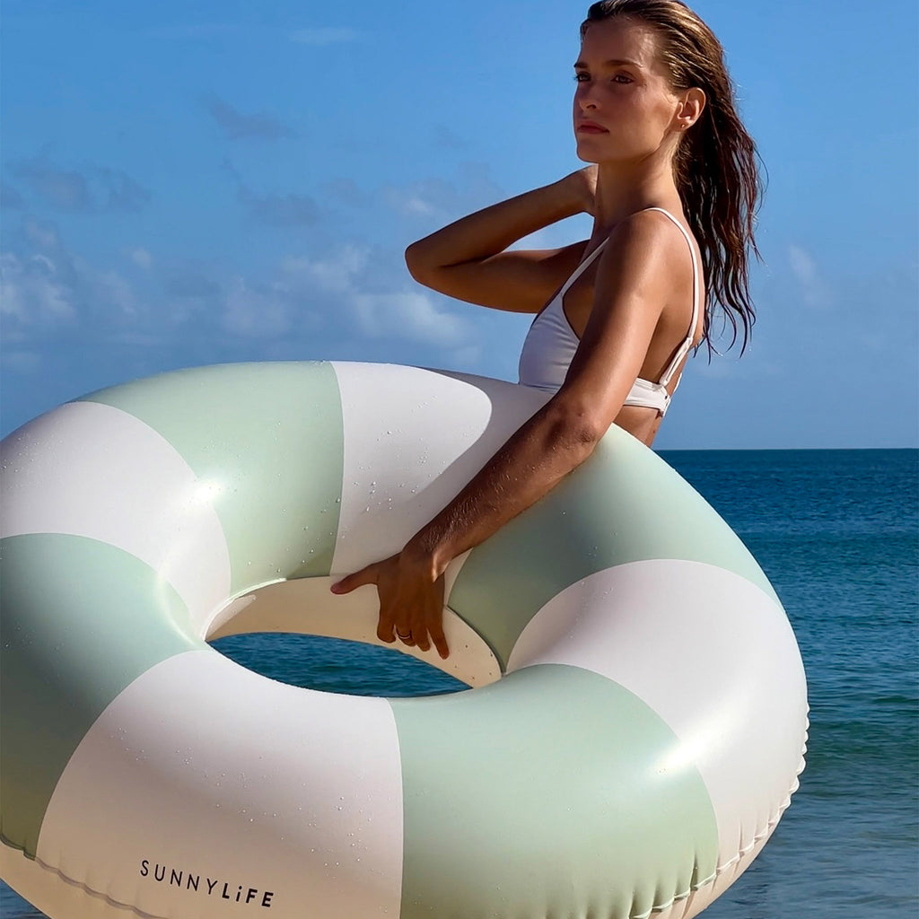 Sunnylife The Vacay Inflatable Tube Pool Ring in soft olive green and white stripes, woman carrying on the beach.