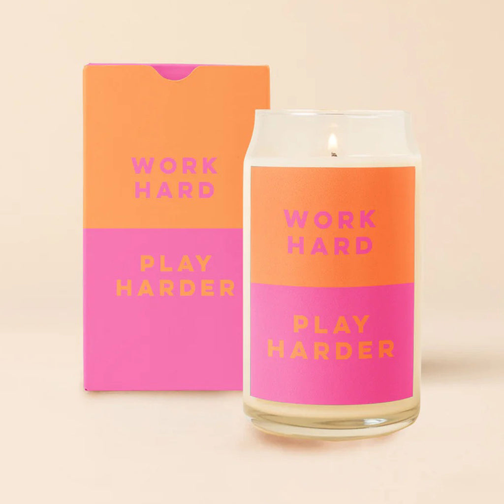 Talking Out of Turn scented soy wax candle in can-shaped glass with orange and bright pink color block label that says "work hard" and "play harder" with gift box that matches the label behind the lit candle.