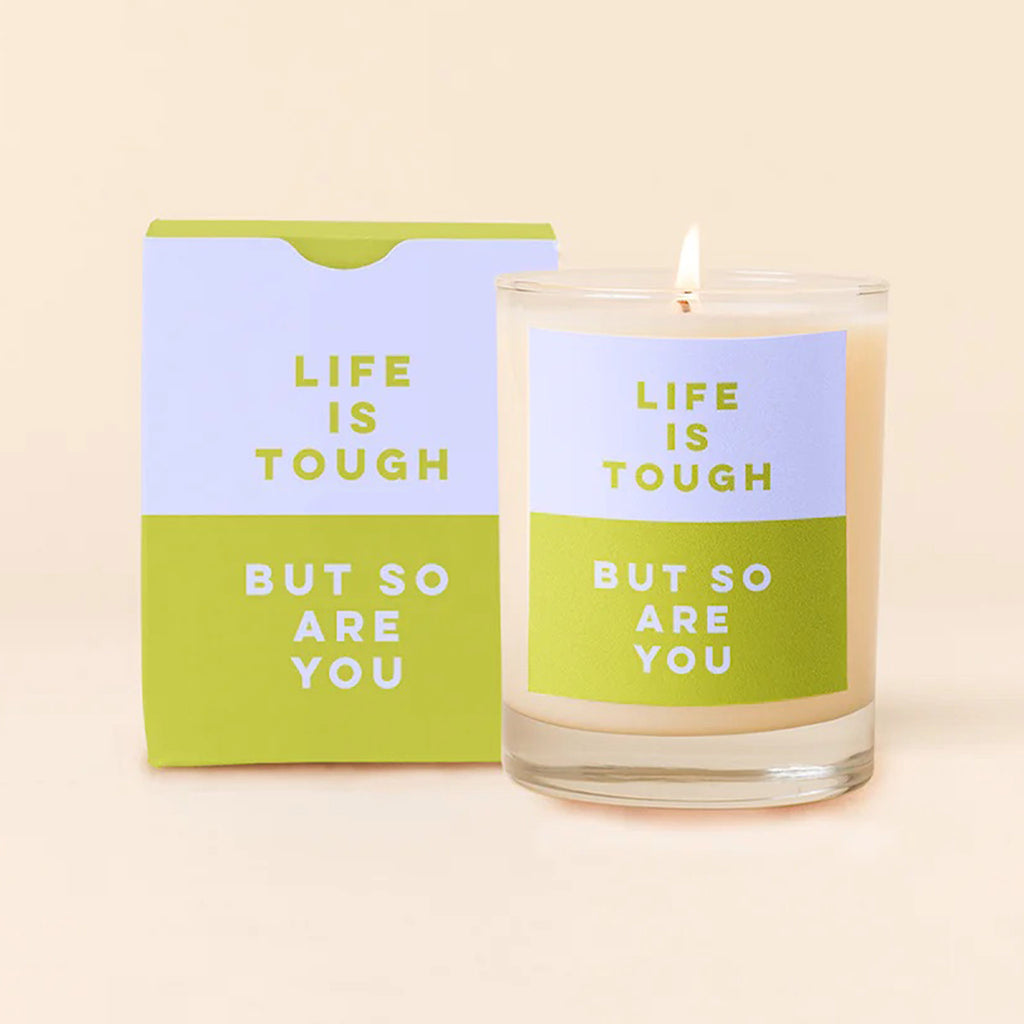 Talking Out of Turn scented soy wax candle in rocks glass with periwinkle and bright olive green color block label that says "life is tough" and "but so are you" with gift box that matches the label behind the lit candle.
