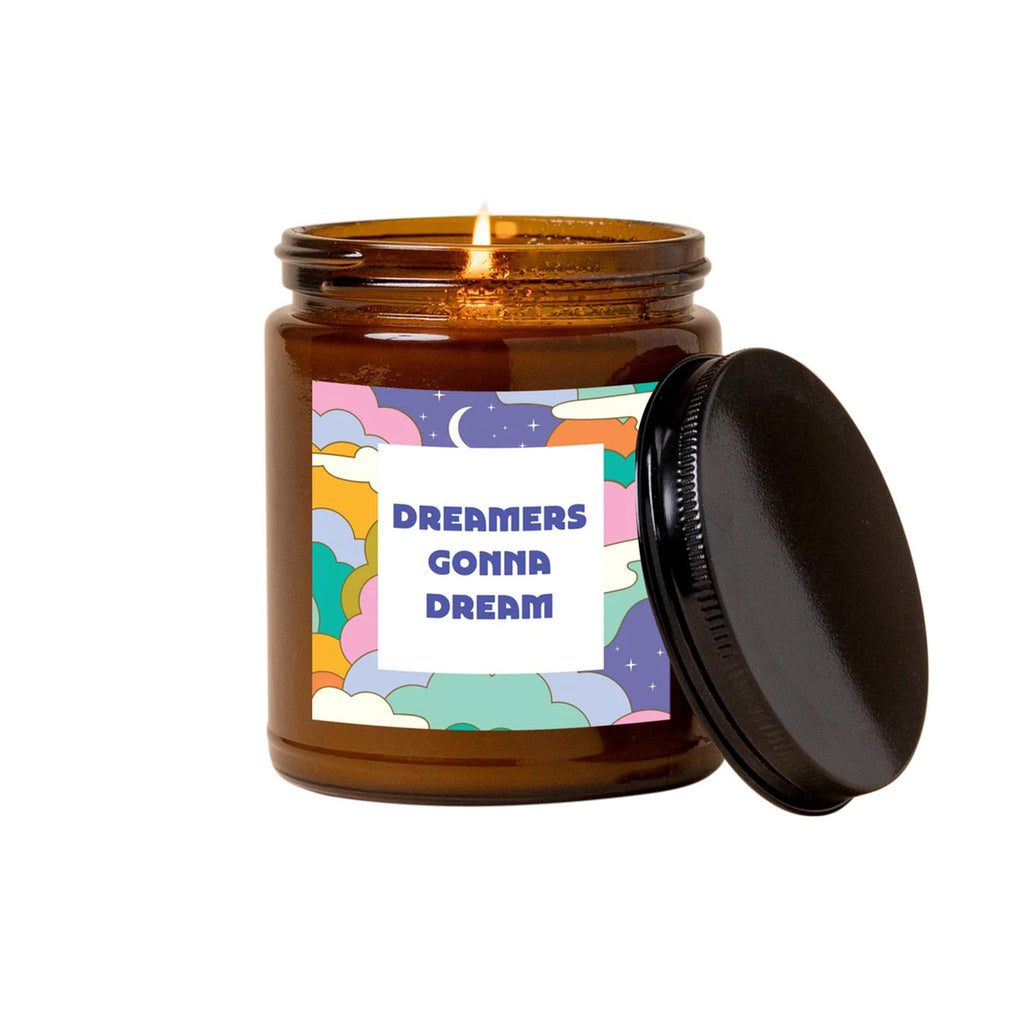 Talking Out of Turn scented candle in amber glass jar with black metallic lid. Label has a colorful cloud illustration with stars and moon and "dreamers gonna dream" in purple lettering.