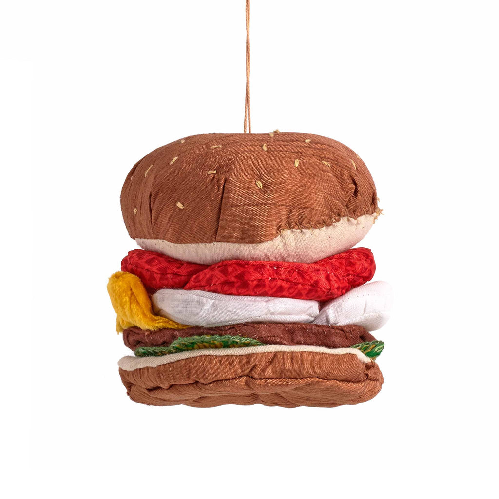 Hamburger-shaped fabric ornament with brown bun, red tomato, white onion, yellow cheese, brown patty, and green lettuce on a white background.