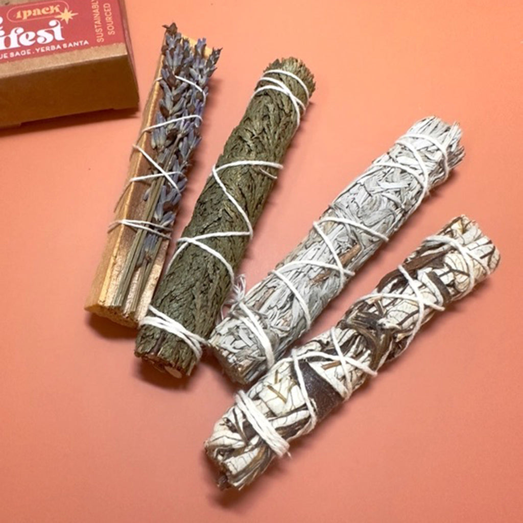 The 12th moon Cleanse and Manifest Kit with lavender palo santo, cedar, blue sage and yerba santa smudge sticks on an orange background.