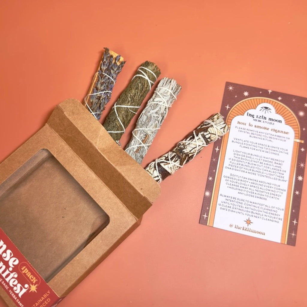 The 12th moon Cleanse and Manifest Kit with lavender palo santo, cedar, blue sage and yerba santa smudge sticks coming out of top of kraft paper box packaging with "how to smoke cleanse" instruction sheet, on orange background.