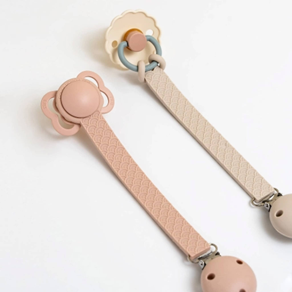 The Cotton Cloud Scallop silicone pacifier holder in ballerina pink and sand, shown with different pacifier types clipped in.