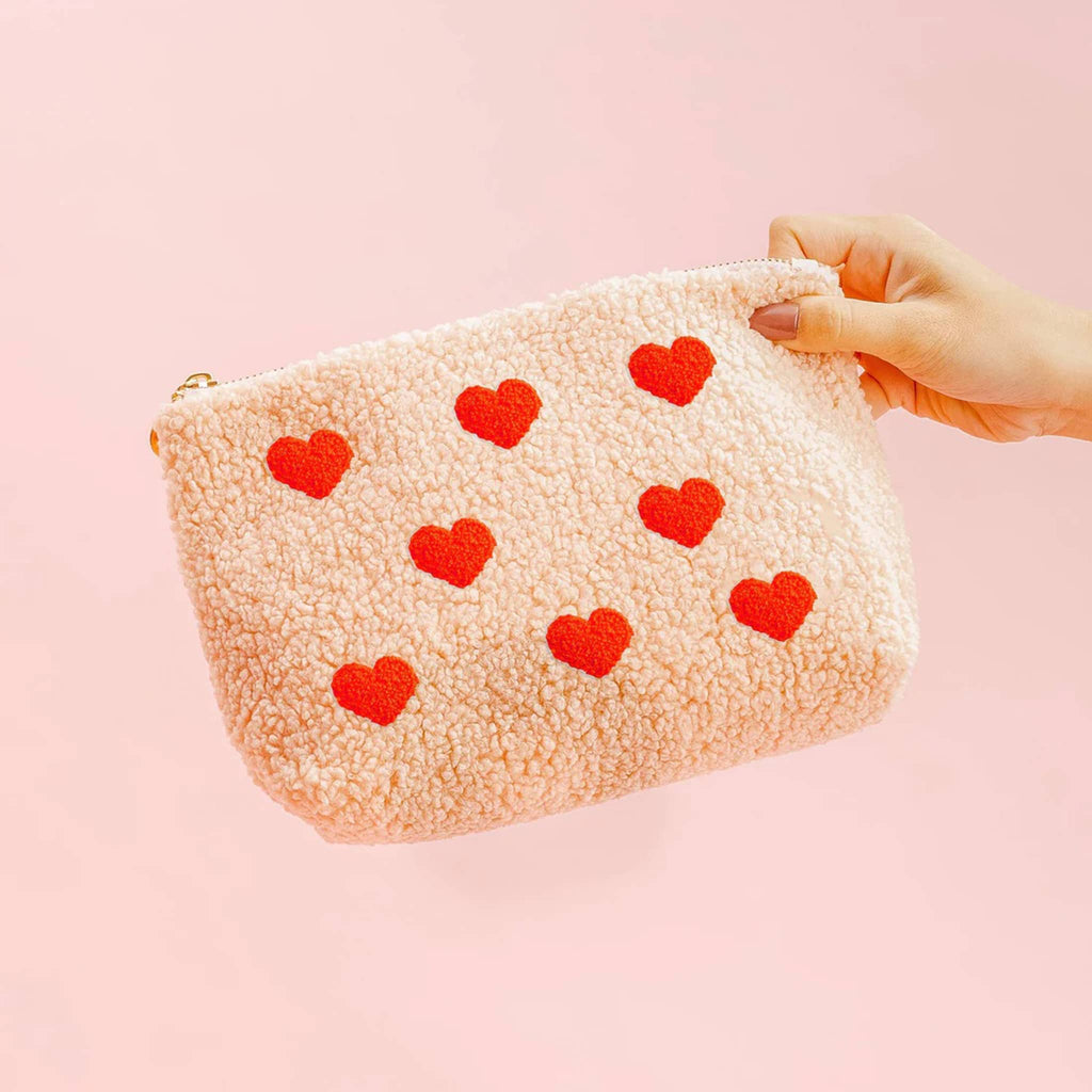 The Darling Effect teddy sherpa zip pouch in pink with red hearts, front view, being held by a hand.