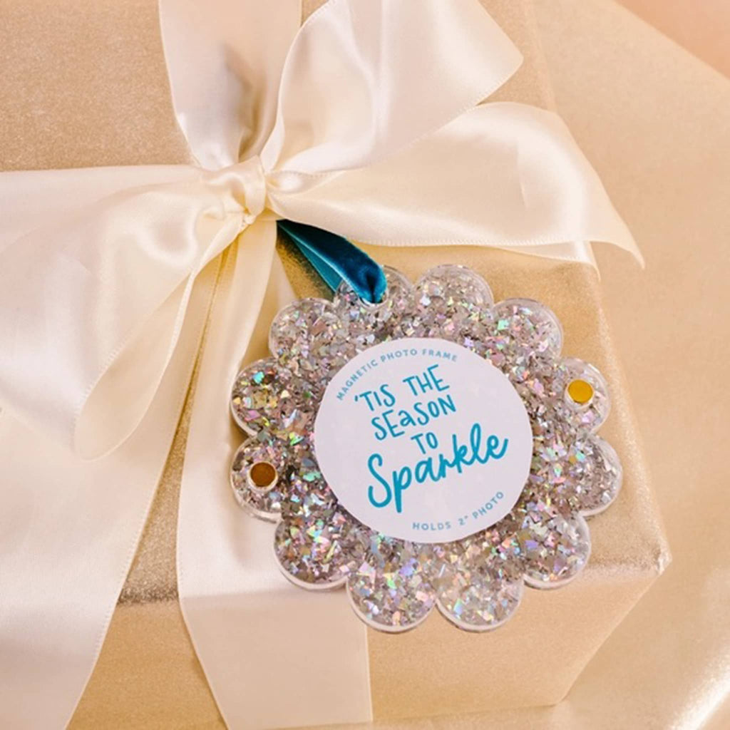 Silver glitter round acrylic magnetic photo frame with scalloped edge and a blue ribbon loop on a wrapped gift box.