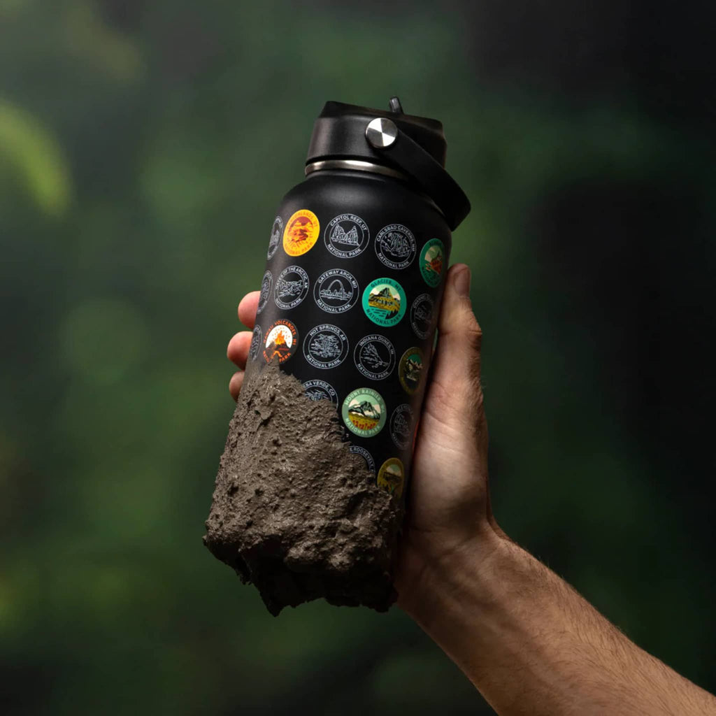 Hand holding a black tumbler with nature-themed stickers and mud on the bottom, against a blurred green background.