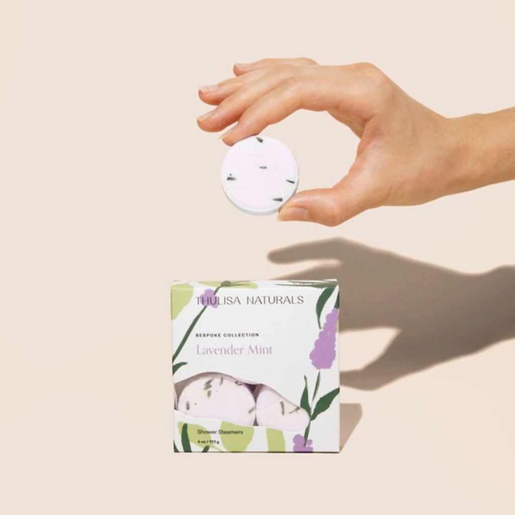 Thulisa Naturals Lavender Mint all natural shower steamers in illustrated box packaging, front view, with a hand holding one of the steamers to show texture.