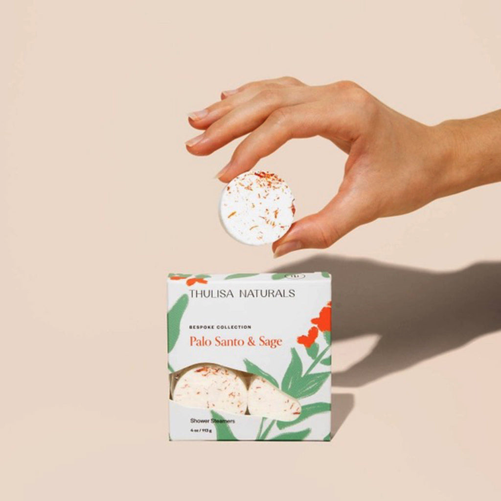 Thulisa Naturals Palo Santo & Sage all natural shower steamers in illustrated box packaging, front view, with a hand holding one of the steamers to show texture.