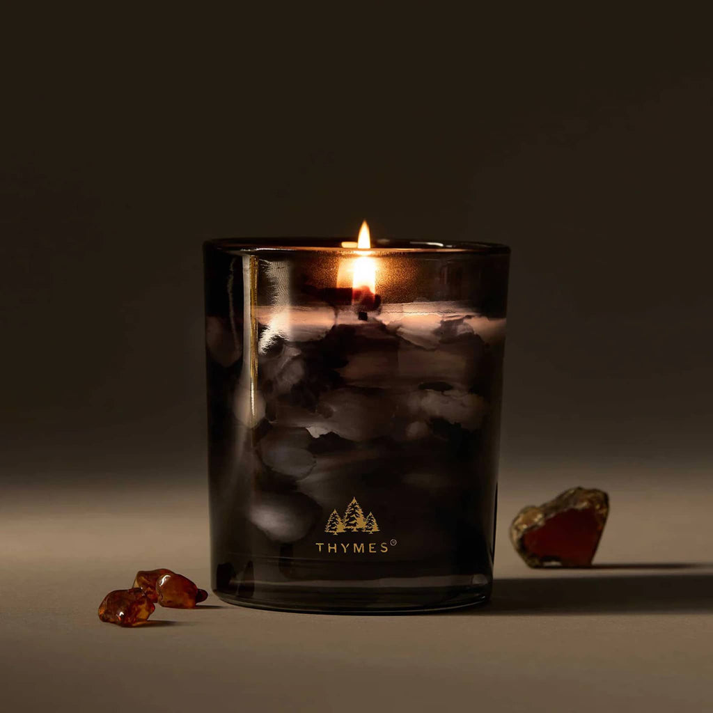 Thymes Noir Woods candle in black marbled glass container and gold logo, wick lit, on a gray background.