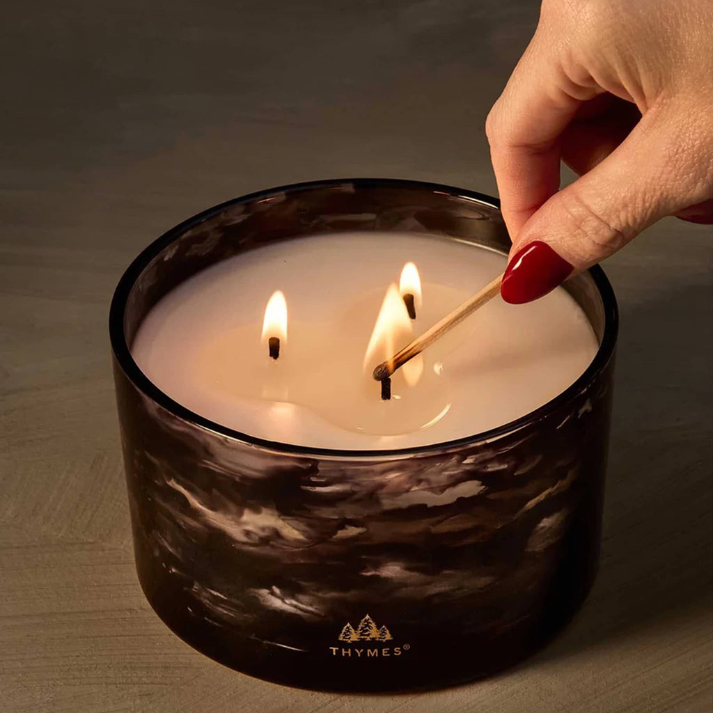 Thymes Noir Woods candle in black marbled glass container with hand lighting the 3 wicks.