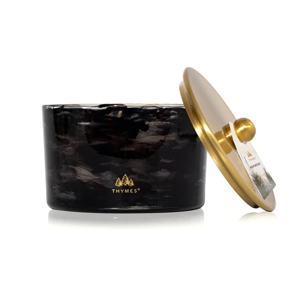 Thymes Noir Woods candle in black marbled glass container with gold lid propped against the side and logo, on a white background.