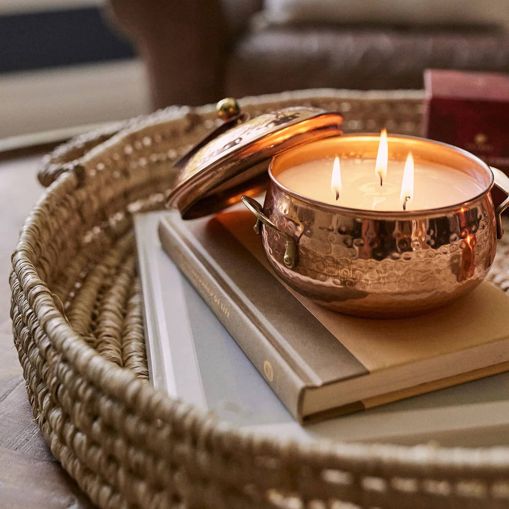 Simmered Cider scented candle in copper pot, lid off and 3 wicks lit, sitting on a book in a woven basket.