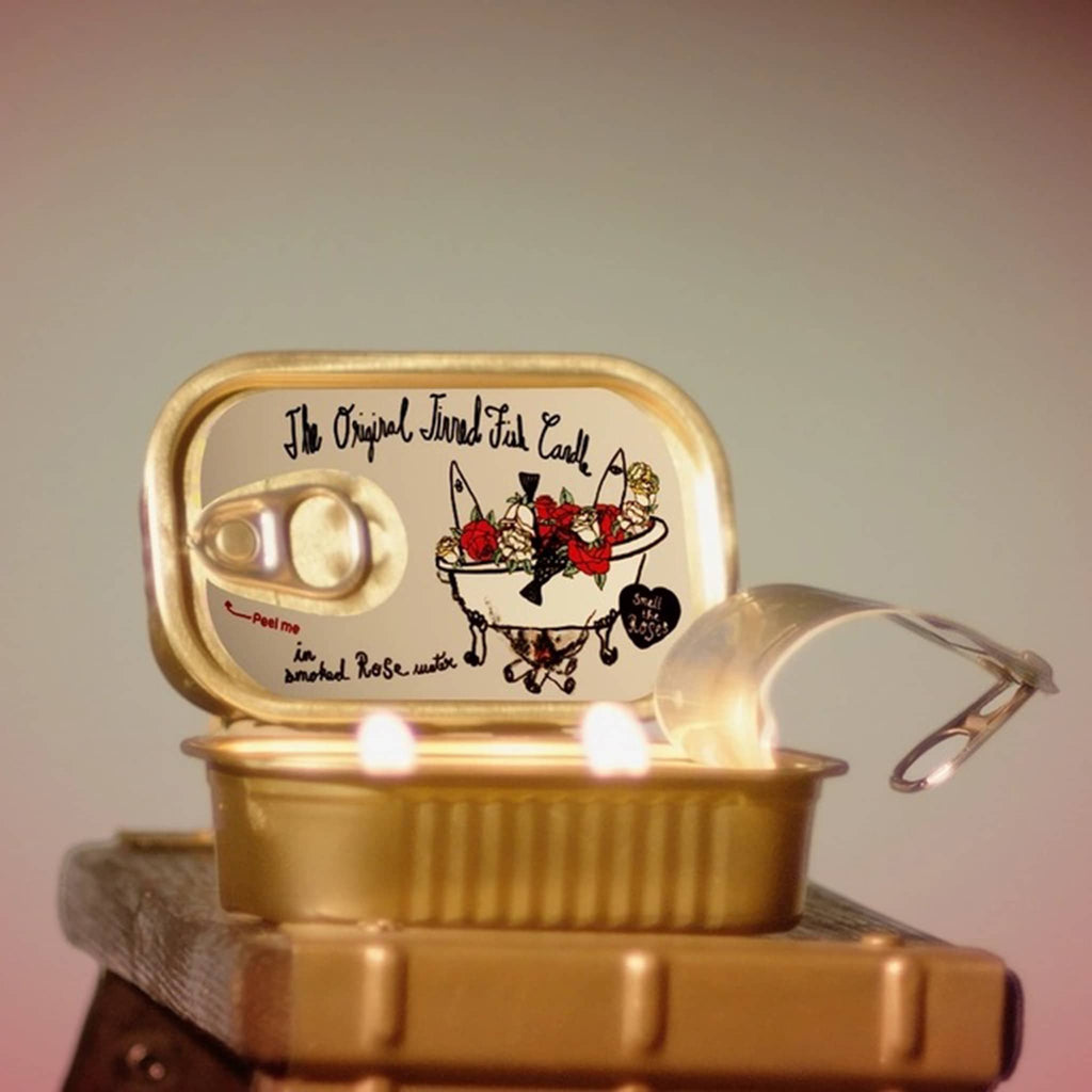 Tinned Fish in smoked rose water, rose scented candle in sardine tin, one lit in front and the other closed to show design on tin lid.
