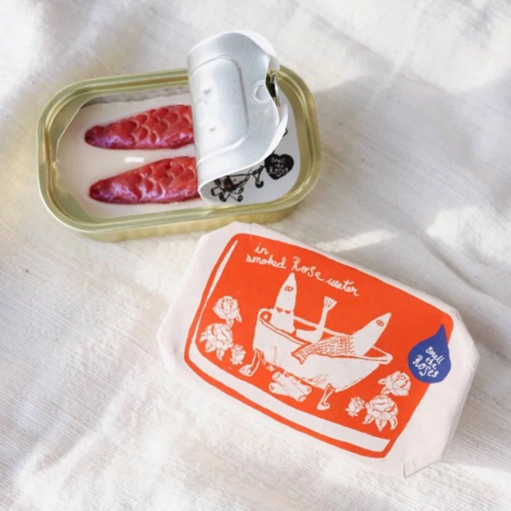 Tinned Fish in smoked rose water, rose scented candle in sardine tin with paper wrap packaging beside an open tin showing red fish in white wax inside.