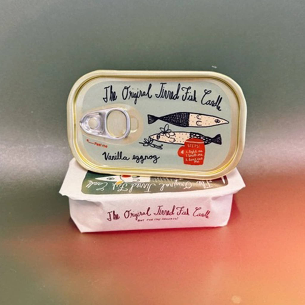 The Original Tinned Fish Candle Vanilla Egg Nog scented holiday soy wax candle in sardine tin packaging sitting on a paper wrapped tin.