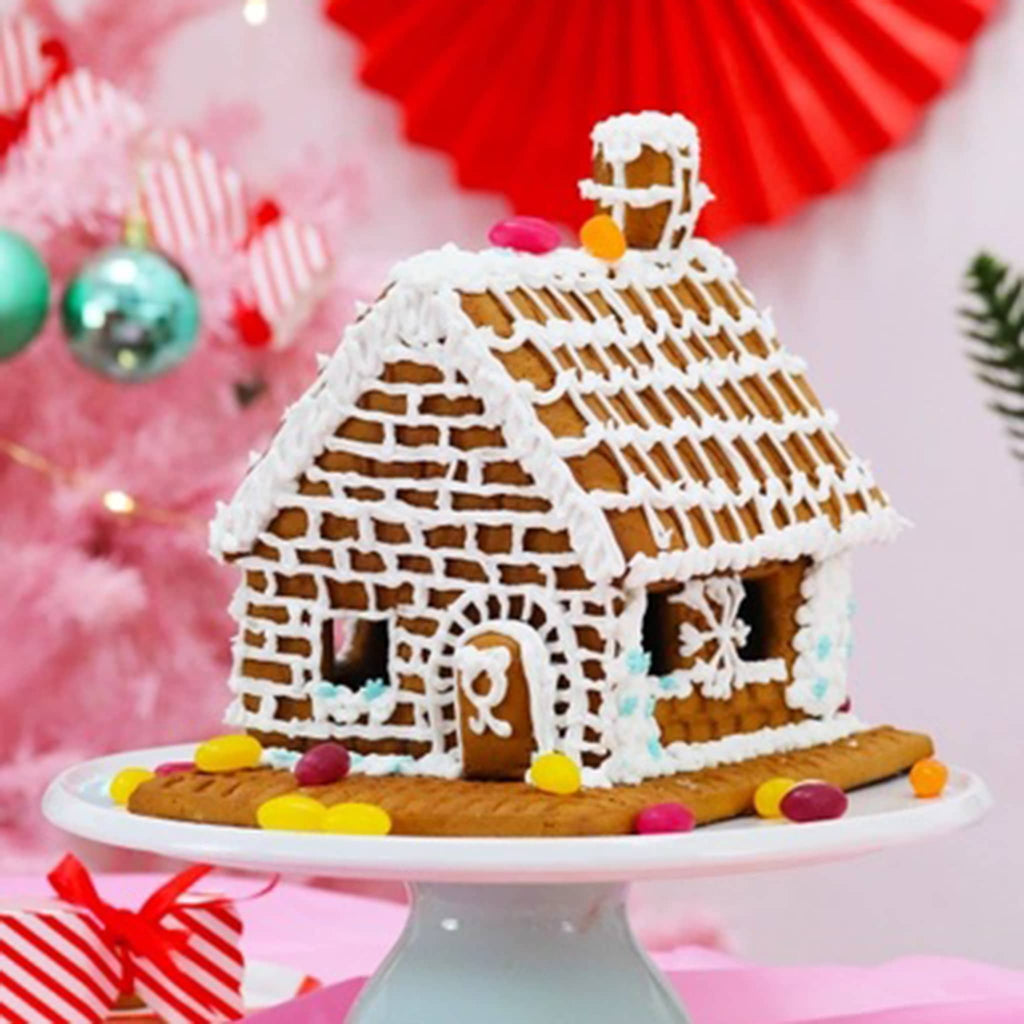 Gingerbread house on a white cake stand with festive decorations in the background.