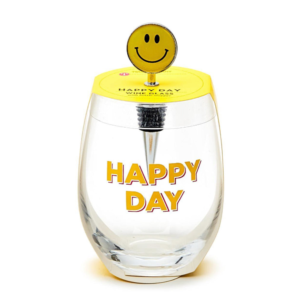 Two's Company clear stemless wine glass with "happy day" in yellow lettering with smiley face bottle stopper, in packaging.