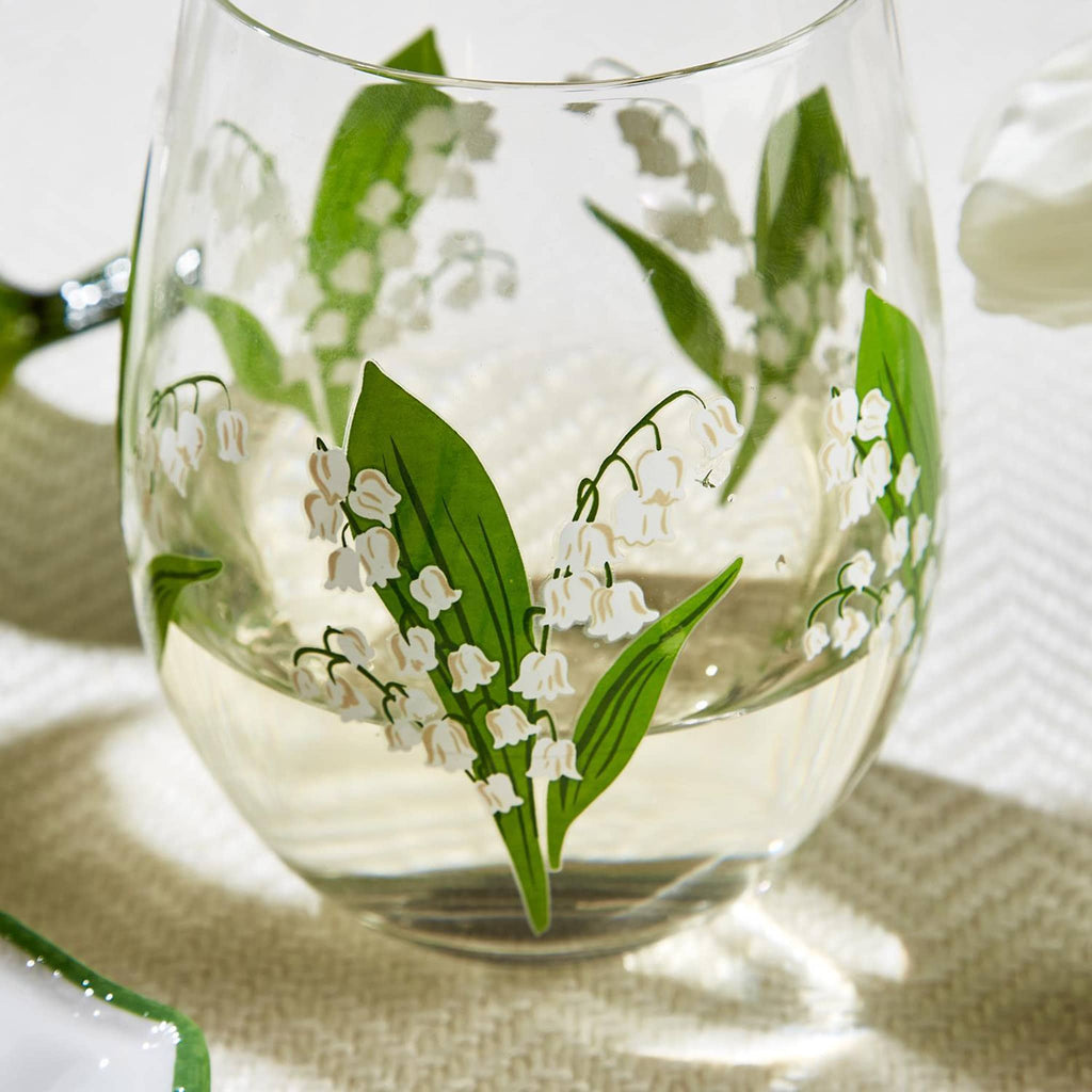 Two's Company clear stemless wine glass with lily of the valley flowers and green leaves all around, filled with white wine on a woven placemat.