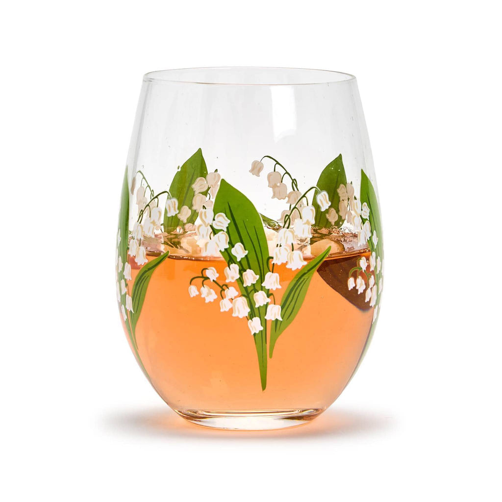 Two's Company clear stemless wine glass with lily of the valley flowers and green leaves all around, filled with orange wine.