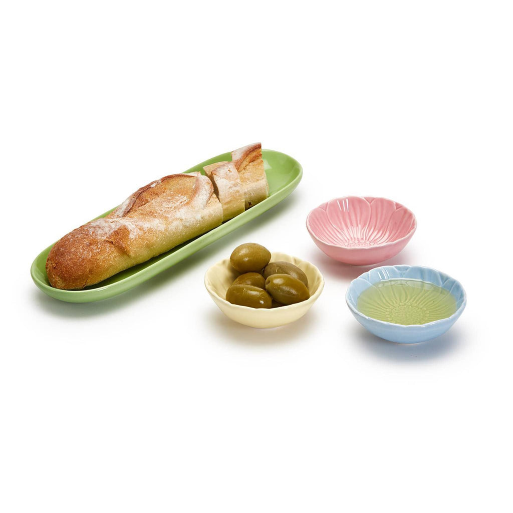 Two's Company pink, blue and yellow ceramic bowls that look like flowers with a green ceramic tray, bowls have olives and olive oil and the tray has a baguette.