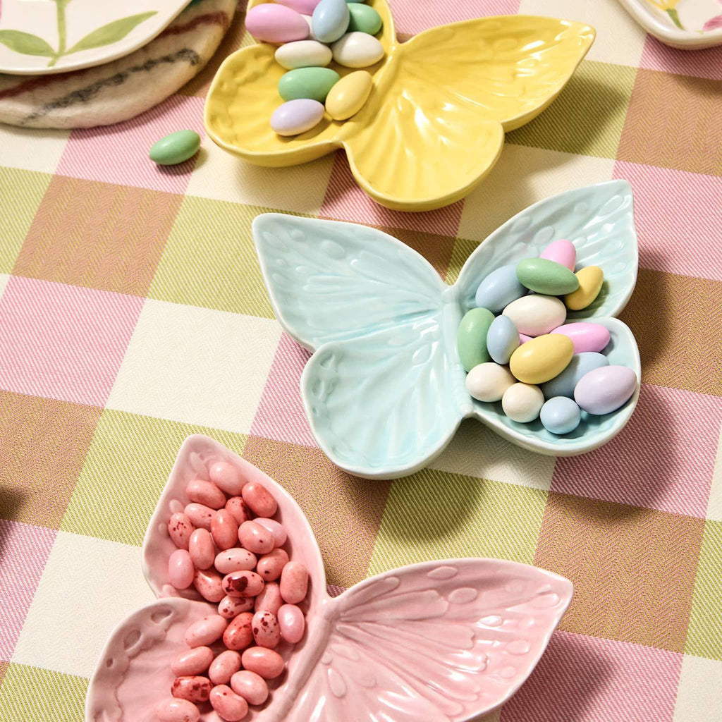 Two's Company Butterfly-shaped porcelain tidbit dish in yellow, pink and blue with candy coated almonds and jelly beans on a gingham tablecloth.