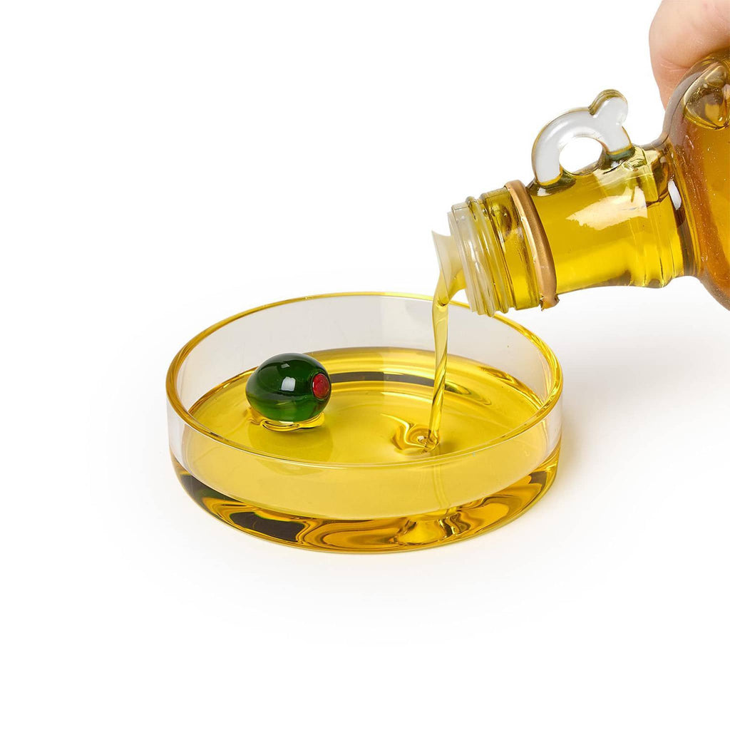 Hand pouring olive oil from a bottle into a small glass dish with a glass green olive inside, on a white background.