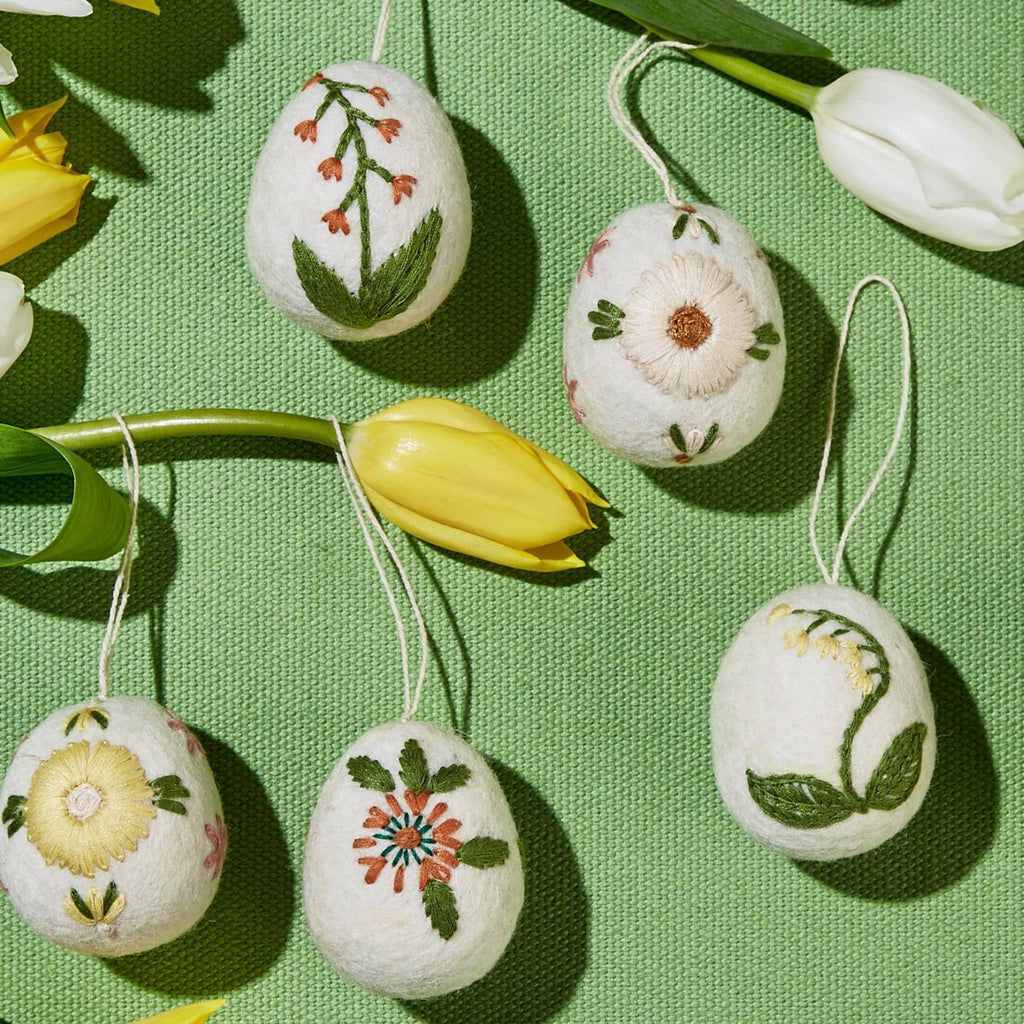Two's Company wool felt eggs with colorful floral hand-embroidery in 5 designs with string for hanging, on a green background with yellow and white tulips.