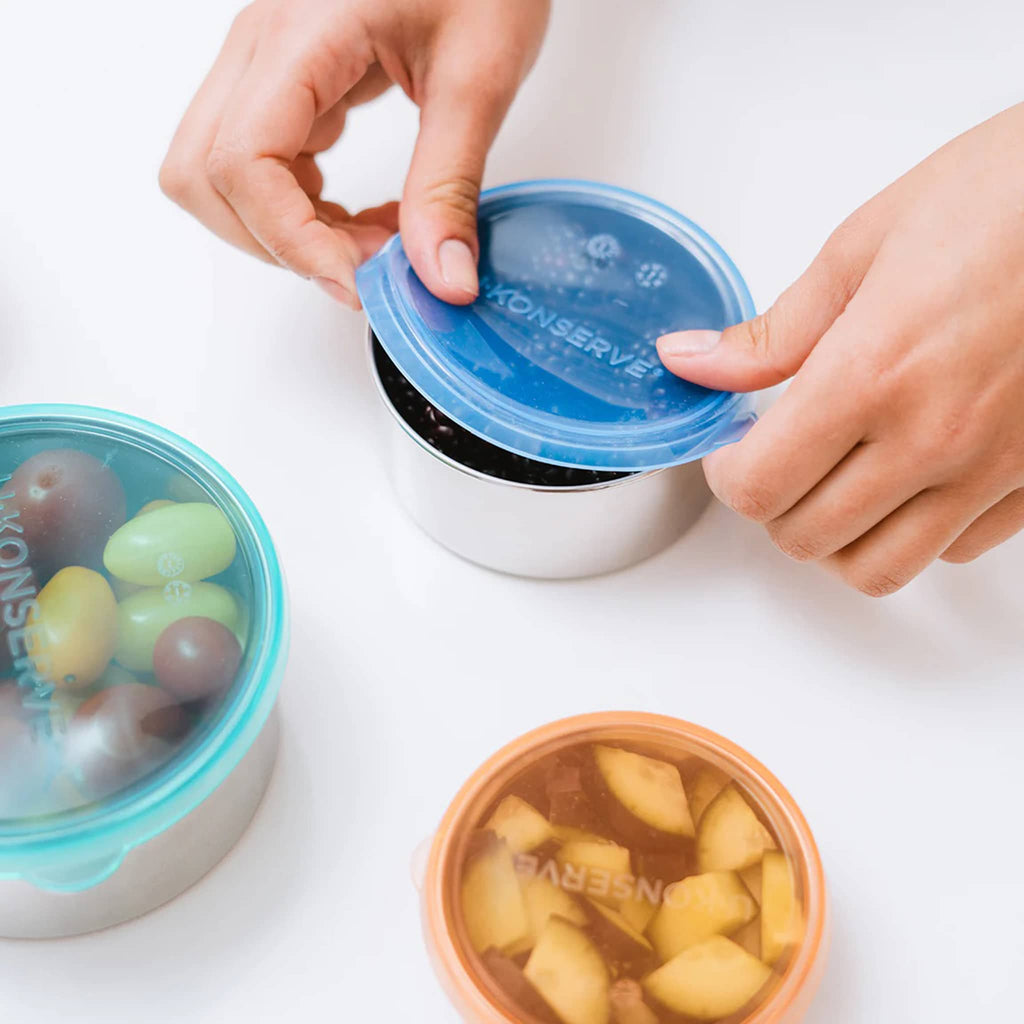 U-Konserve set of three round stainless steel food containers in 5 ounce, 9 ounce and 16 ounce capacity, each with a plastic-free, leakproof, silicone lid in orange, blue or turquoise. Containers are shown with fruit inside with hands removing one of the lids.