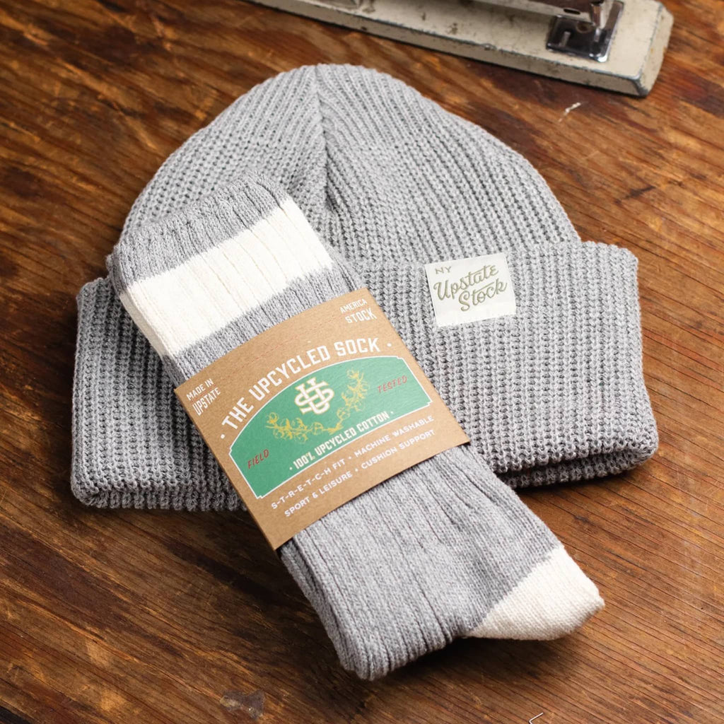 Gray and white sock with 'The Upcycled Sock' packaging and matching beanie, on a wooden surface.