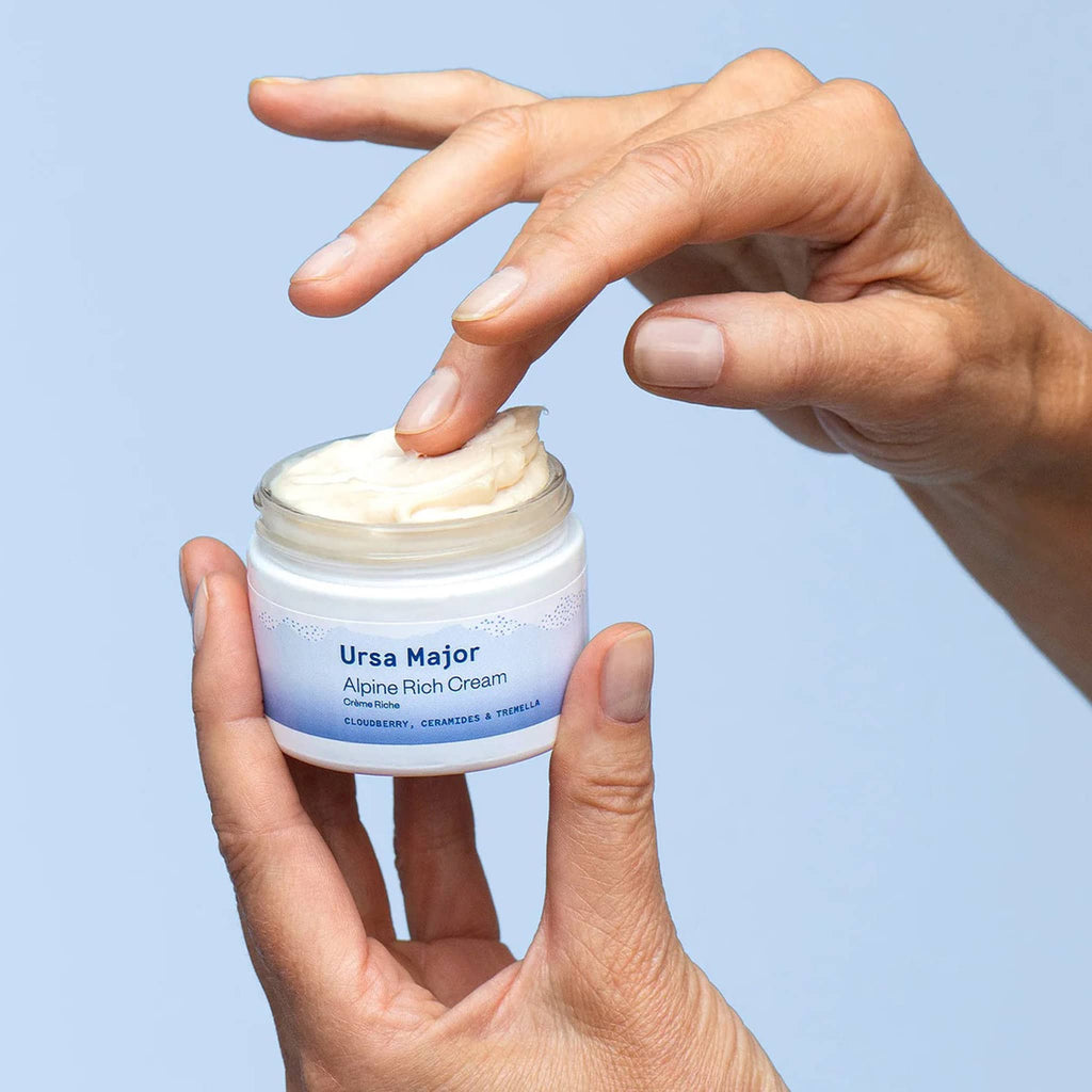 Ursa Major Alpine Rich Cream facial moisturizer in glass tub with lid off to show texture, being held between fingers with other hand in cream.