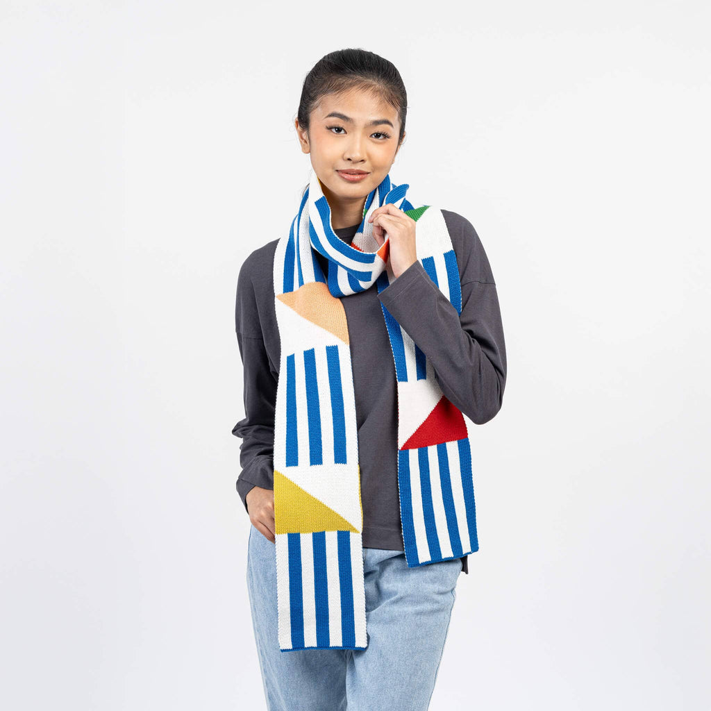 Blue and white striped knitted skinny scarf with sailing flag-inspired colorful insets, on a person with a gray shirt and jeans.