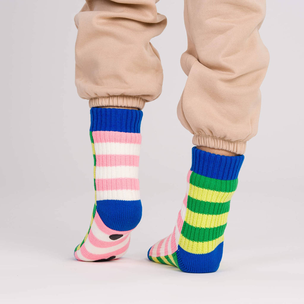 Verloop Super Stripe pink, green and blue slipper house socks worn with beige sweatpants, on a gray background.