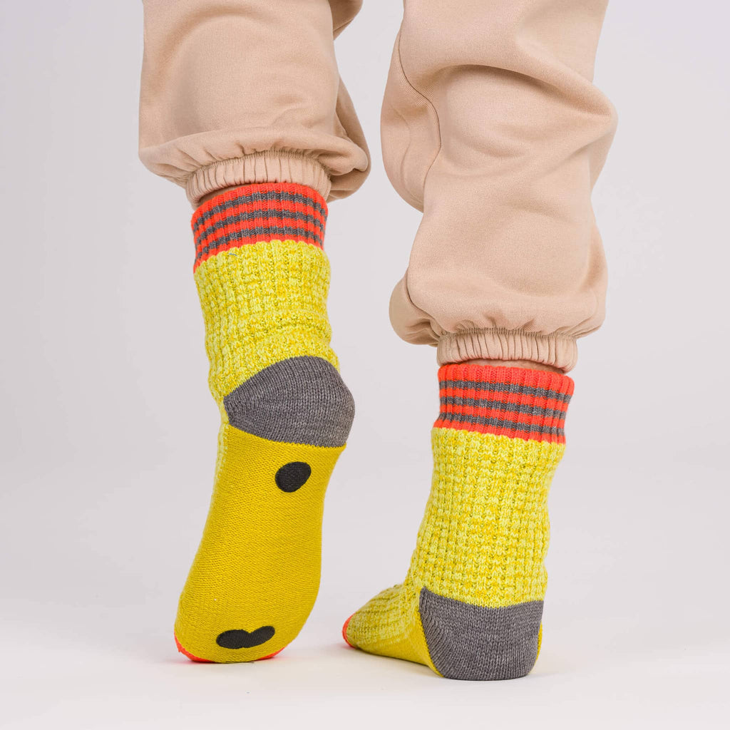 Verloop Varsity Knit yellow, orange and gray color-blocked slipper house socks worn with beige sweatpants, on a gray background.
