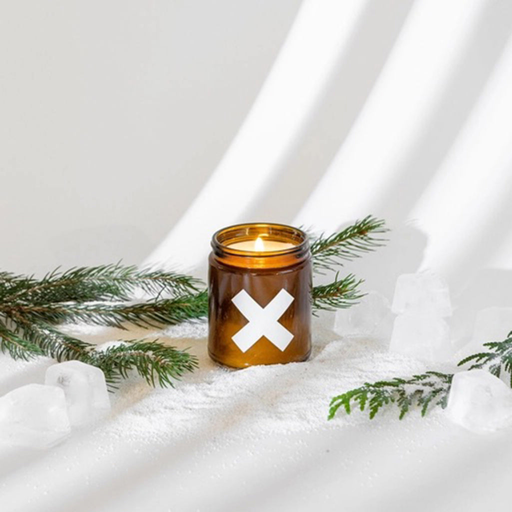 Wax Buffalo 9 ounce evergreen scented soy wax candle in amber glass jar with white X on the front, wick is lit and candle has evergreen branches, ice cubes and fake snow around it on a white background.
