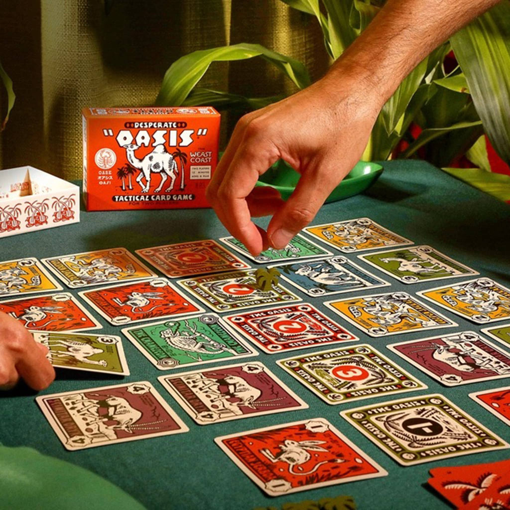 2 people playing a card game with 'Desperate Oasis' box in the background.