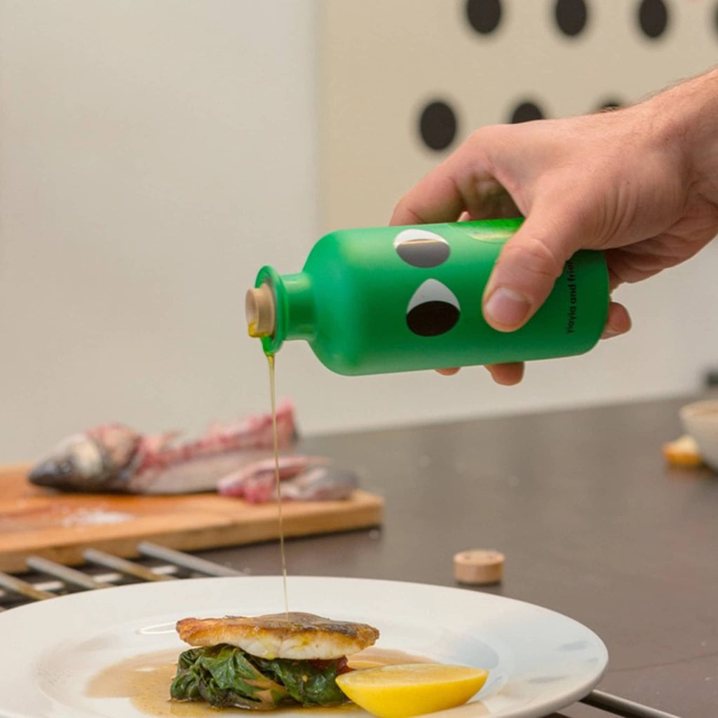 yiayia and friends basil infused greek extra virgin olive oil in green glass bottle with eyes, being poured onto a fish dish.