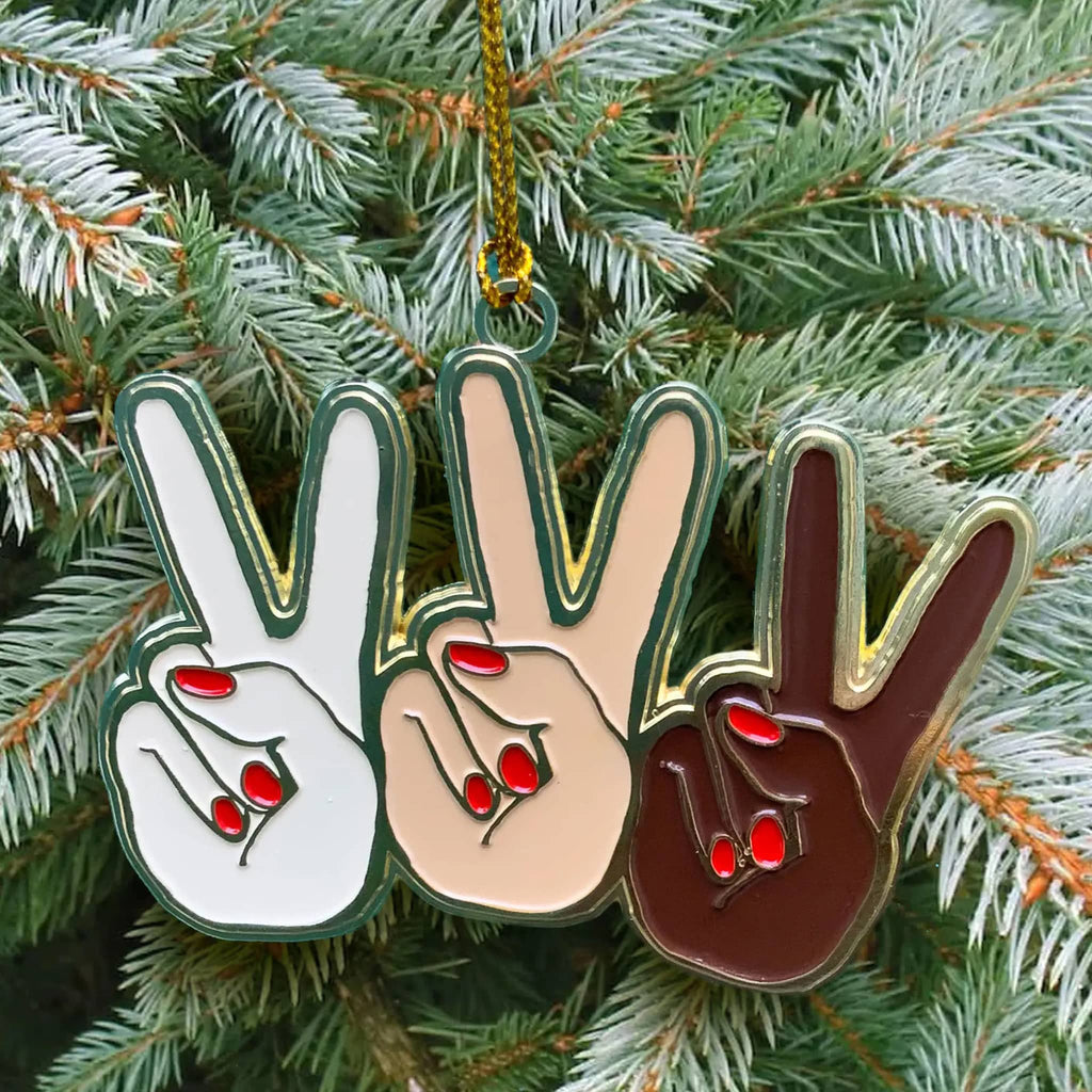 Front view of an enamel holiday ornament that features 3 hands in different skin tones flashing a peace sign, ornament is hanging on an evergreen tree.