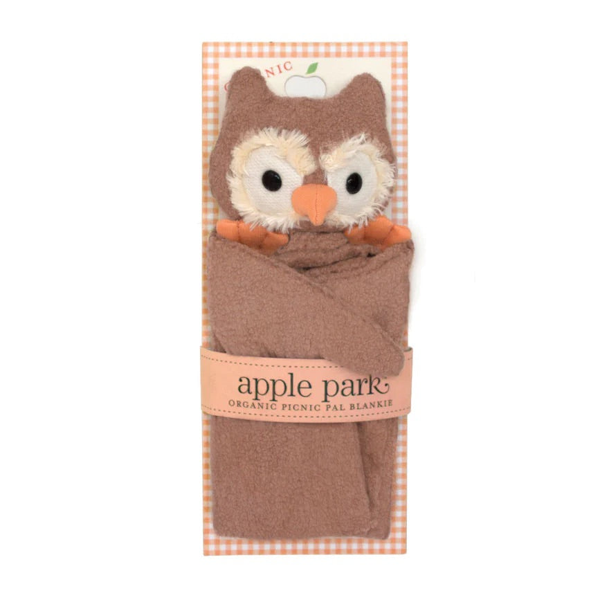 Brown baby blanket with an owl head, black button eyes surrounded by white circles and an orange beak and feet. Held onto a paper card with peach gingham print by a peach belly band that says "apple park, organic picnic pal blankie".