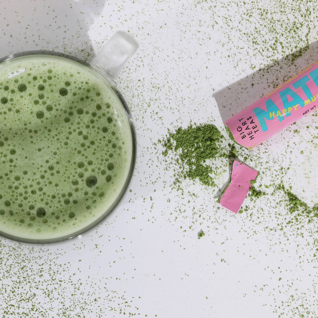 Big heart company happy matcha green tea single serving stick packet open with tea powder spilled onto white background next to a clear glass mug filled with matcha tea.