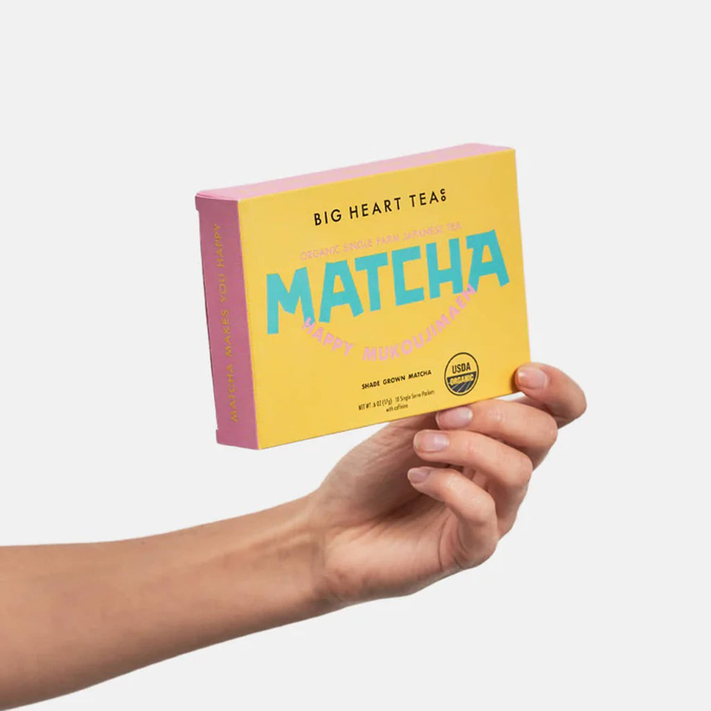 Big heart company happy matcha green tea single serving stick packets packaged in a golden yellow and pink box, being held in the palm of an outstretched hand.