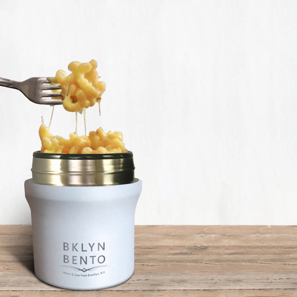White stainless steel insulated food jar on a wood surface, filled with macaroni and cheese with a silver fork scooping out a bite.