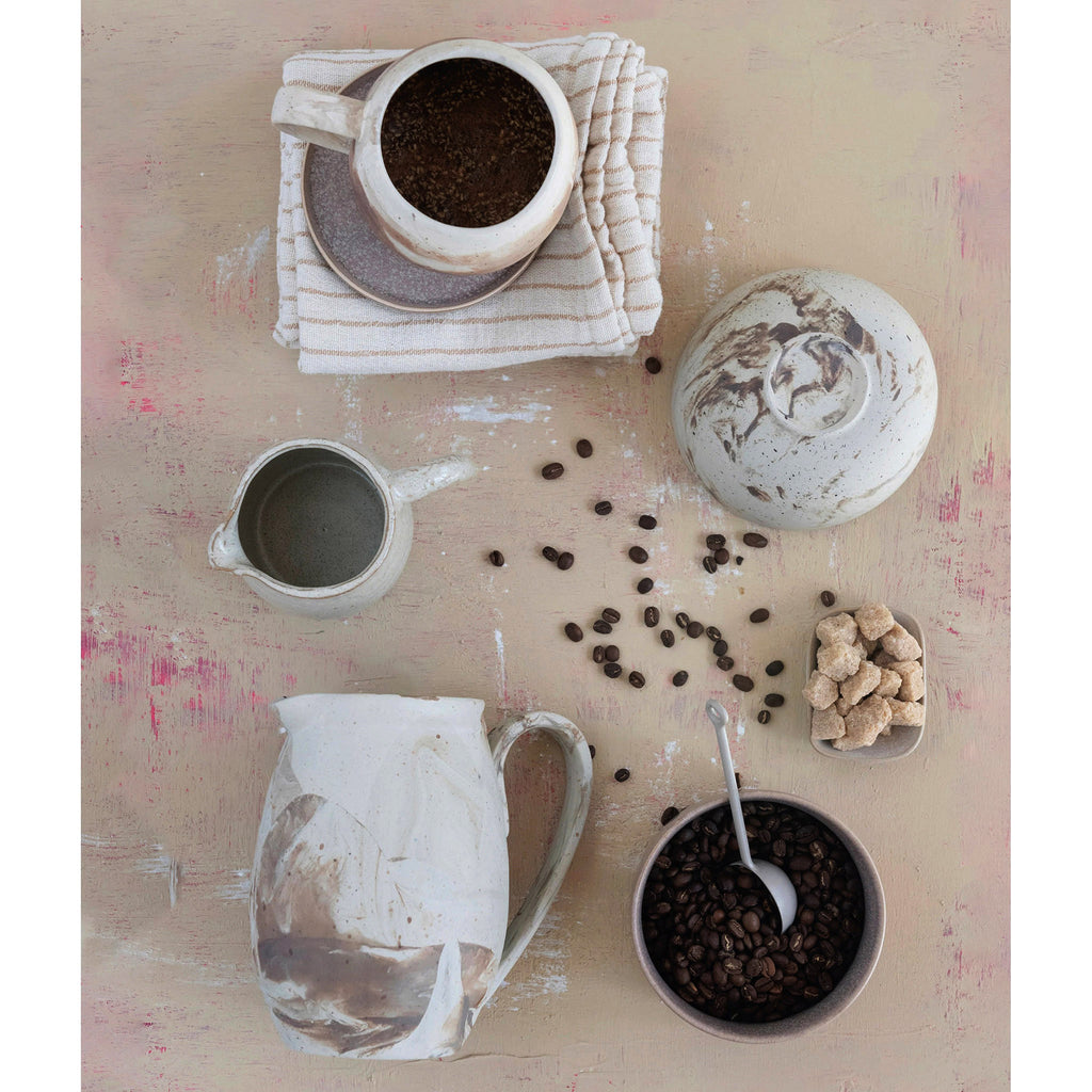 Bloomingville sand colored stoneware bowl, mug and pitcher with brown reactive glaze in swirl patterns. Propped with coffee beans and raw sugar cubes.