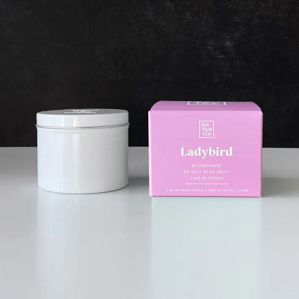 Botanica Ladybird scented candle in white travel tin with lid beside a pink and white gift box.