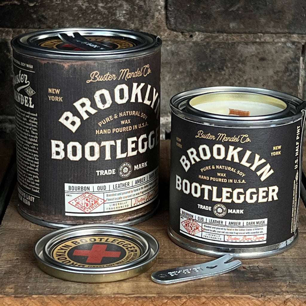 Buster Mandel Co. Brooklyn Bootlegger scented soy wax candle in pint and half pint metal tins with lid and black label with vintage look. Candles are on a wood surface with aged brick in background, small candle has lid off to show wood wick.